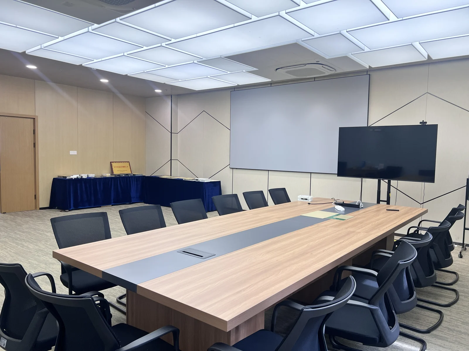 会议长桌、投影幕布和显示屏，后方可见蓝色桌布展示台